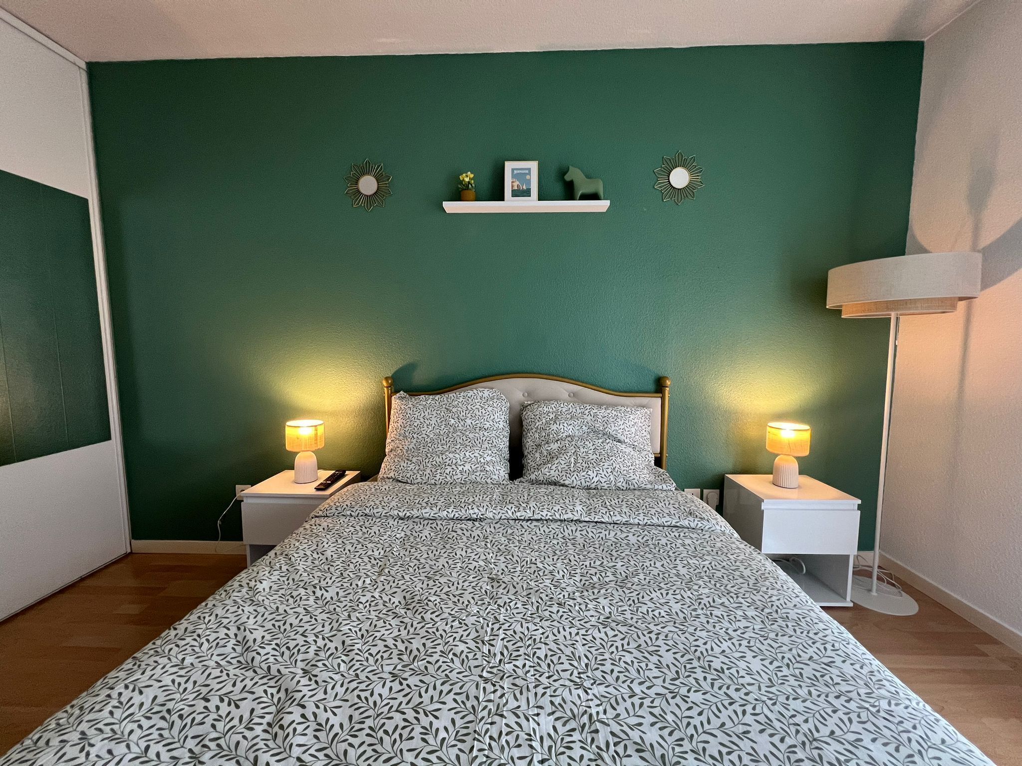 A bedroom with a bed, two bedside lamps, a nightstand, a wall-mounted shelf with decorative items, and a wall clock.