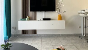 Un salon moderne avec un téléviseur à écran plat posé sur une console blanche, une horloge au mur et deux tables rondes ornées de plantes en pot.