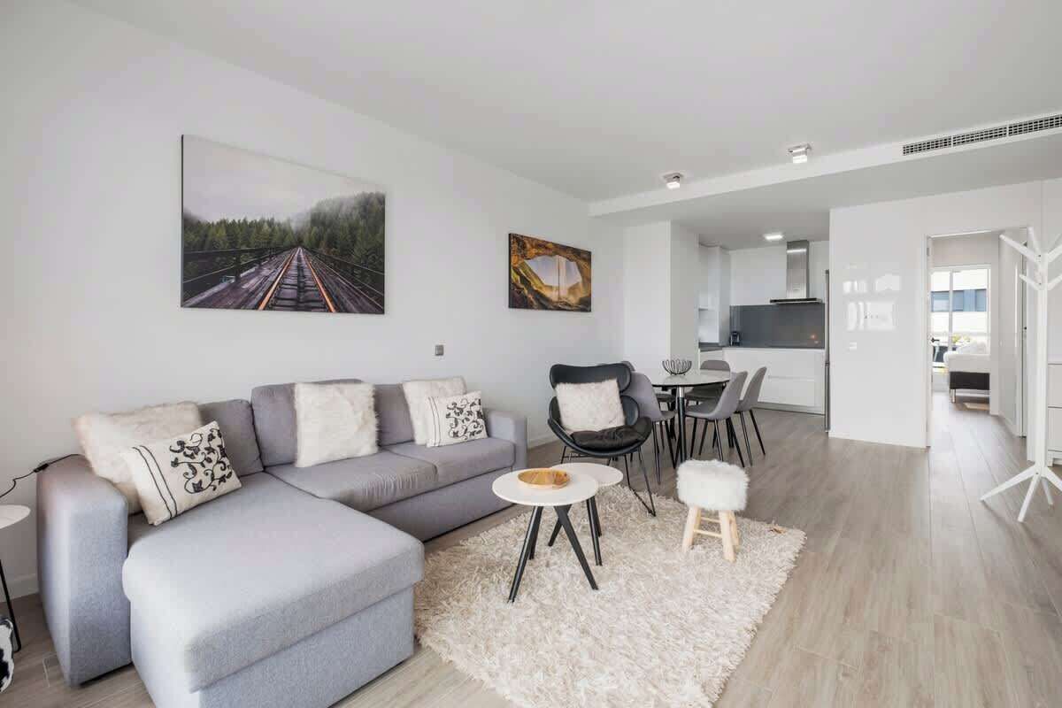Una sala de estar moderna con un sofá seccional gris, una mesa de centro redonda y un alfombra mullido blanco. Hay una zona de comedor con una mesa pequeña y sillas, y una cocina al fondo.