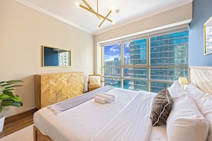 A modern hotel room with a large bed, a wooden herringbone cabinet, a view of the city through large windows, and a ceiling light fixture.