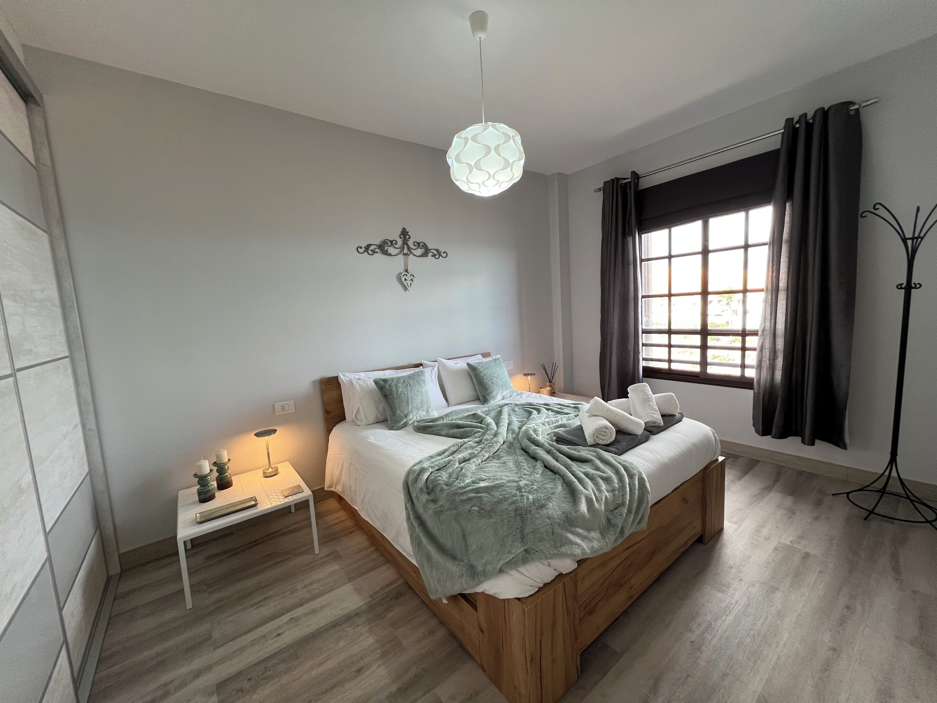 A modern bedroom with a wooden bed, a bedside table with a lamp, and a view of the outdoors through a window with curtains.