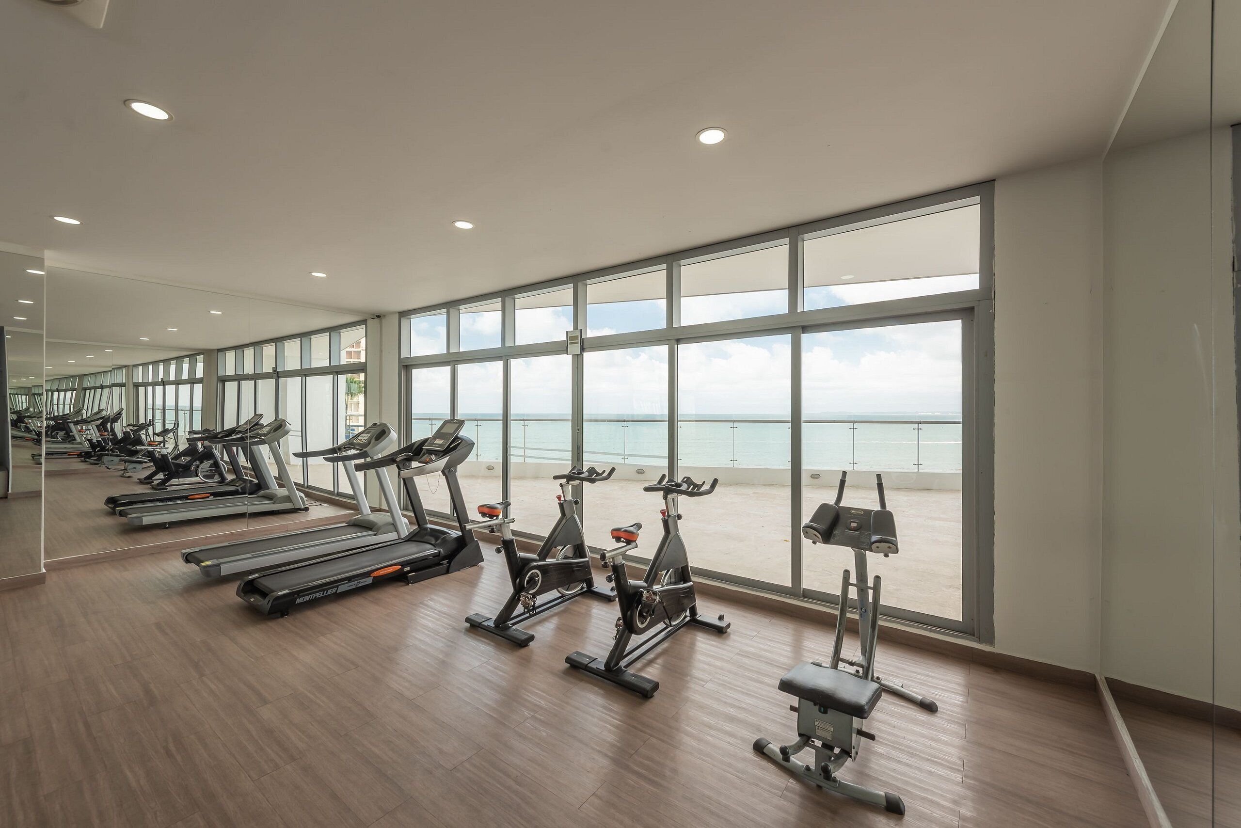 Een fitnessruimte met loopbanden, hometrainers en een roeimachine, grote ramen met uitzicht op zee, en een houten vloer.
