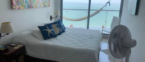 A hotel room with a bed, a hammock, a fan, a painting, and a view of the sea.