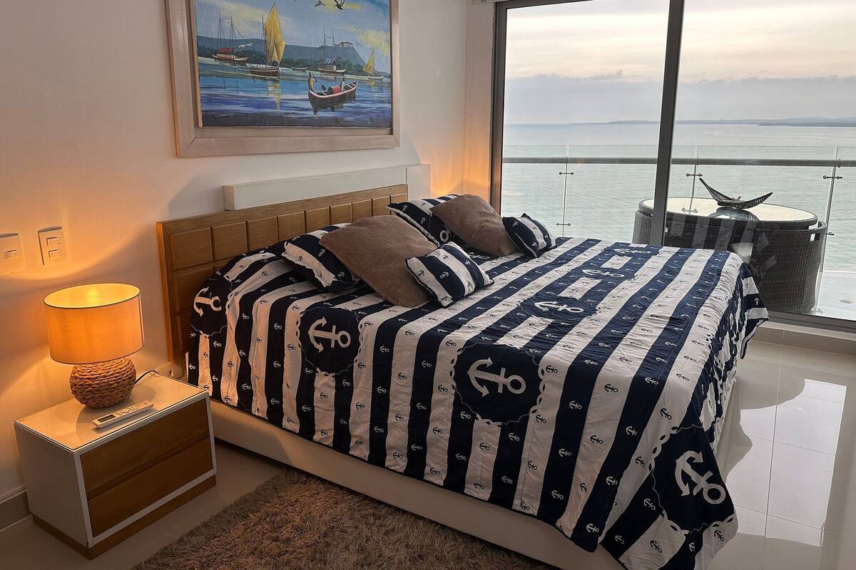 Een slaapkamer met een bed met gestreepte beddengoed, een nachtkastje met een lamp, een schilderij van een zeegezicht en een balkon met uitzicht op de zee.