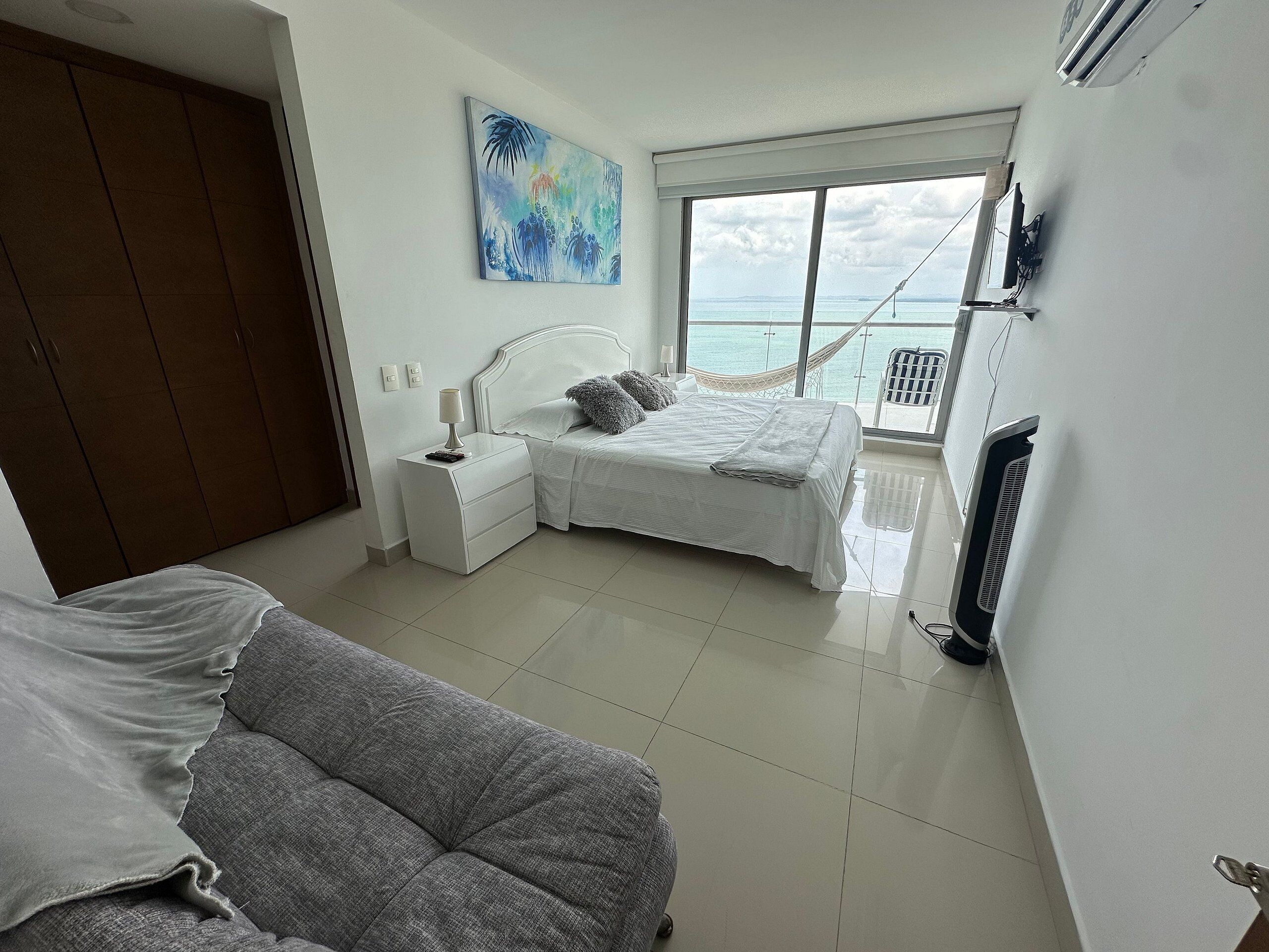A bedroom with a bed, nightstands, a white wardrobe, a grey sofa, and a large window with a view of the sea.