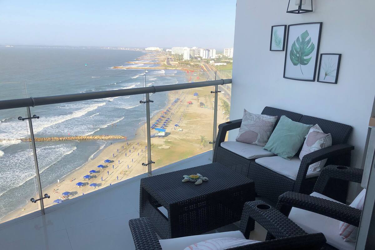 Een balkon met rieten meubilair, uitzicht op het strand en ingelijste bladeren aan de wand.
