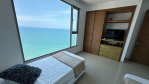 A bedroom with a large window overlooking the sea, a bed with a white mattress and a blue pillow, a wooden wardrobe, and a TV stand with electronic devices. - Edificio Alta Mar de Marbella (Cartagena)