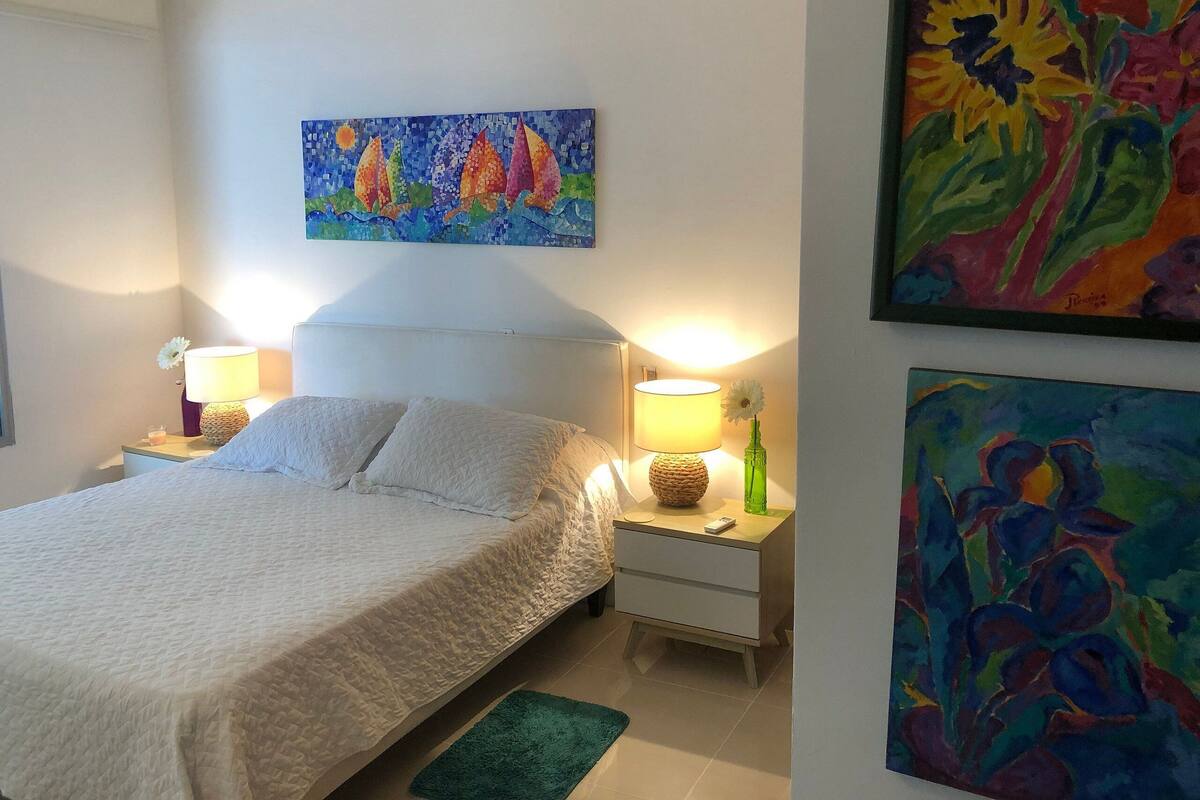 Een slaapkamer met een bed, twee nachtkastjes met lampen, een bijzettafeltje met een kaars, en twee schilderijen aan de muur.