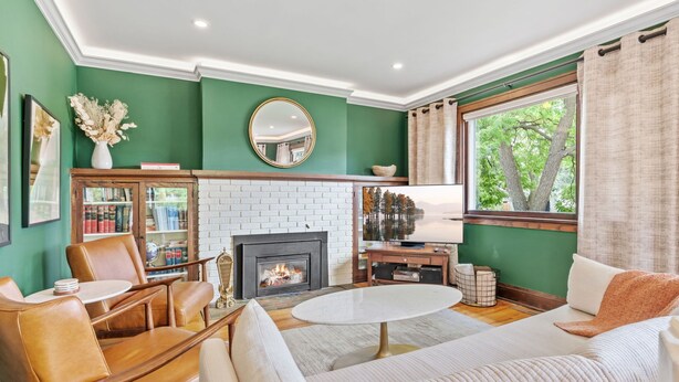 Een woonkamer met een open haard, groene wanden, leren fauteuils, een witte bank en een groot raam met gordijnen.
