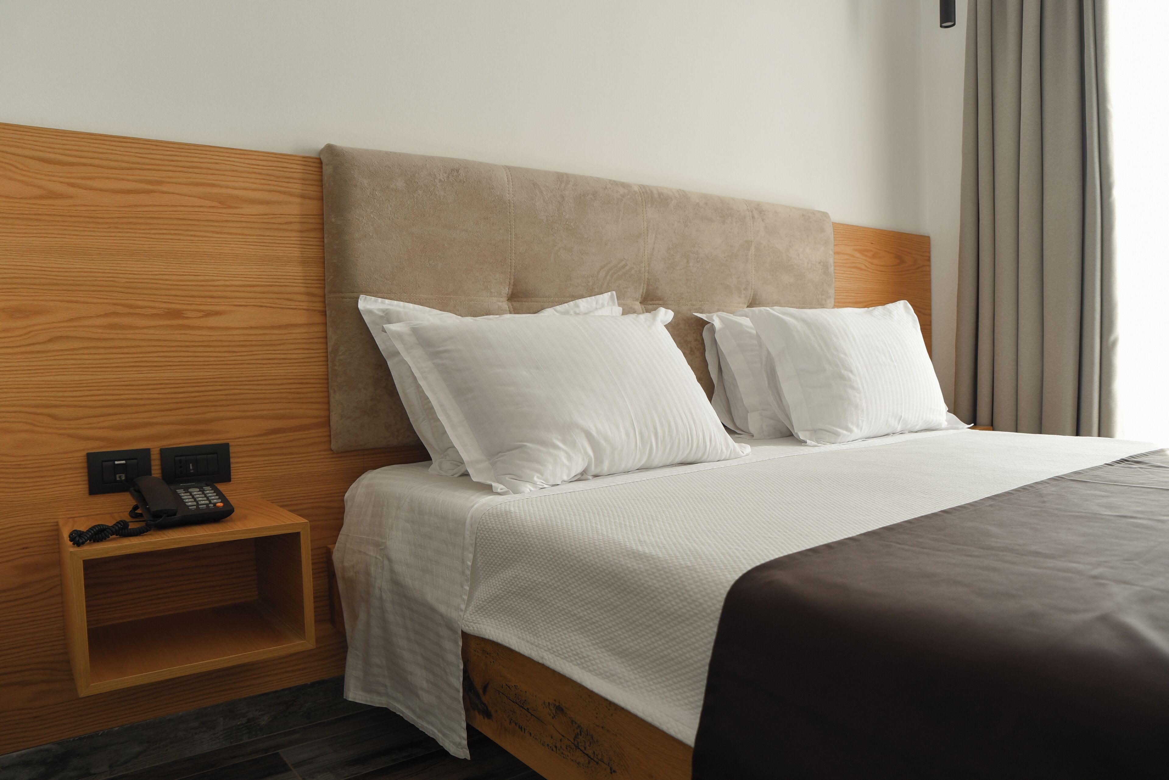A neatly made bed with white bedding and a wooden headboard, a bedside table with a phone, and a wall-mounted light switch.