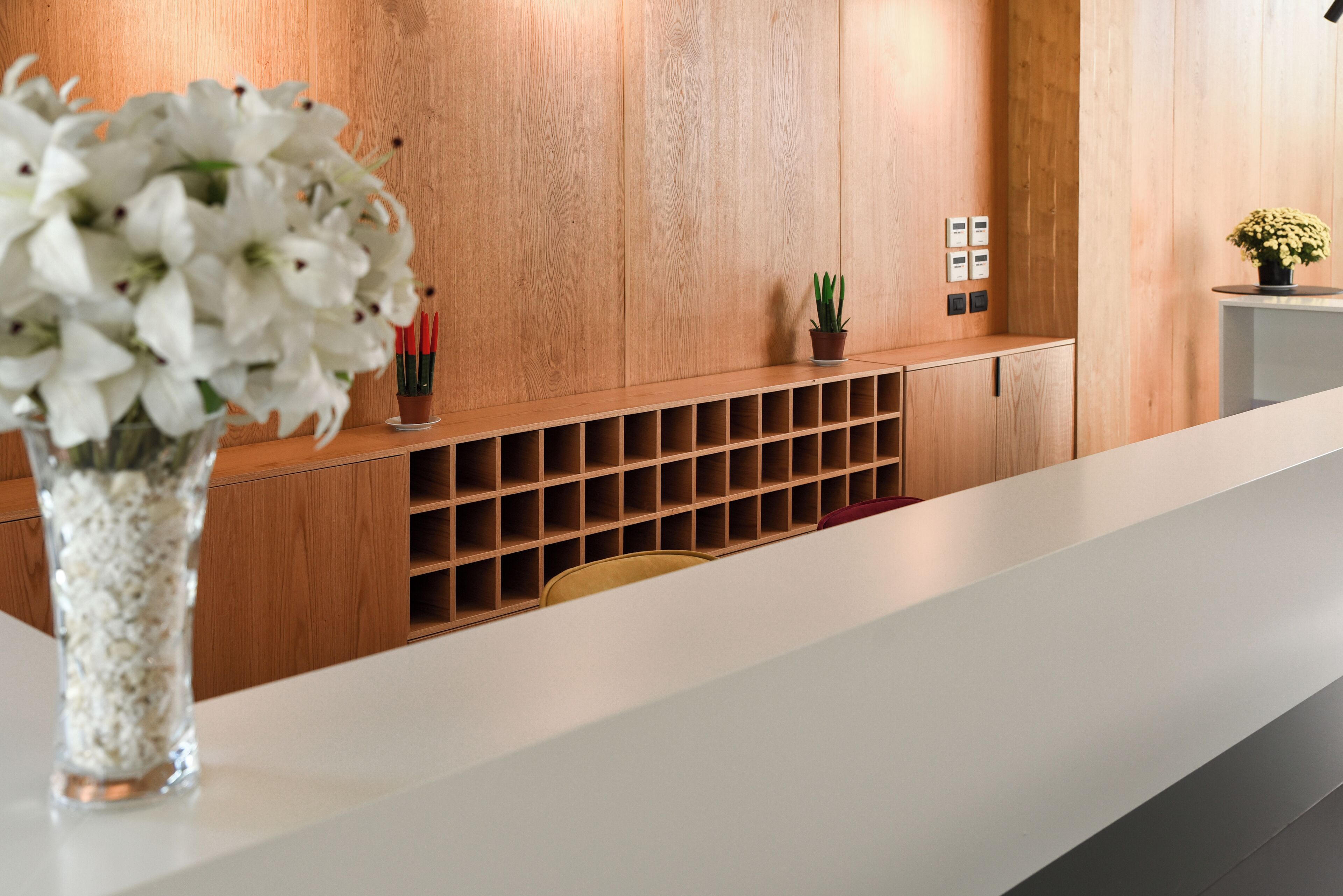 Un área de recepción con pared de paneles de madera, un mostrador blanco, un jarrón con flores blancas, una pequeña planta en maceta y una estantería con velas rojas.