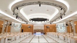 A large, well-lit conference room with a stage, rows of white tables and chairs, chandeliers, and a modern ceiling design. - Hotel Albulena (Mamurras)