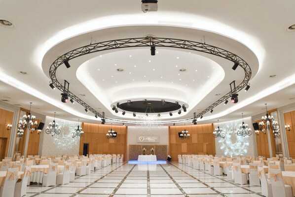 A large, well-lit conference room with a stage, rows of white tables and chairs, chandeliers, and a modern ceiling design.