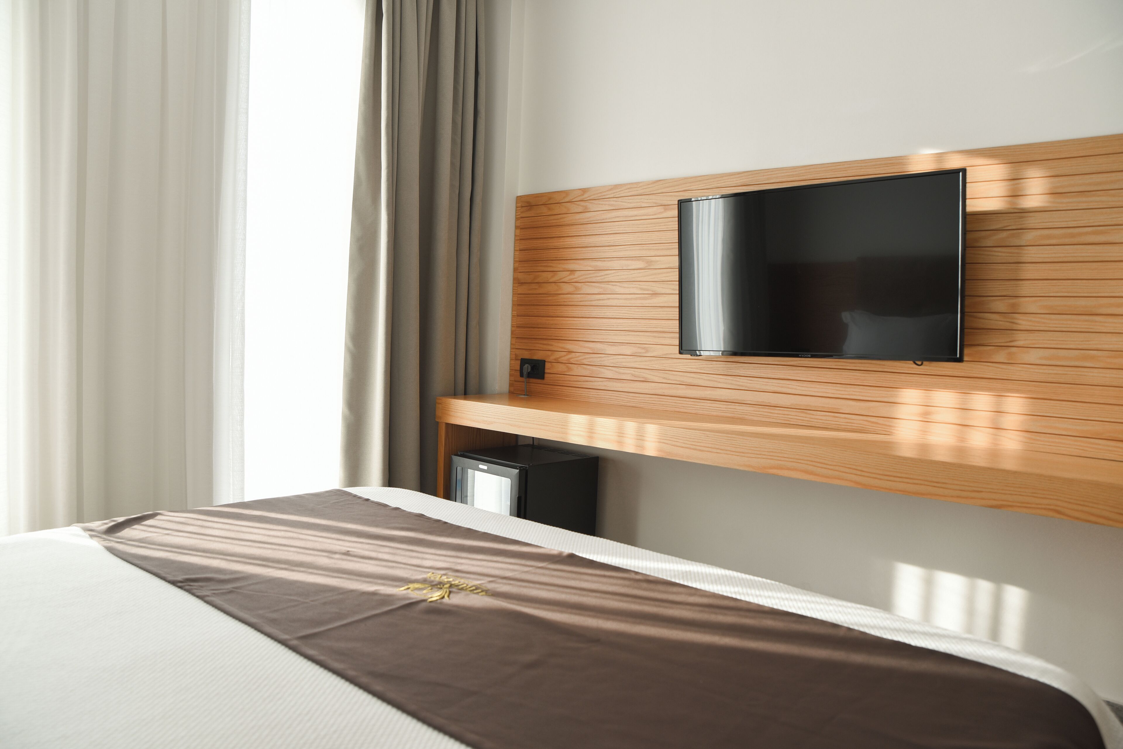 Moderni hotellihuone, jossa on puinen sängynpääty, siihen kiinnitetty litteänäyttöinen televisio, ruskealla päiväpeitteellä varustettu sänky ja pieni minibaari.