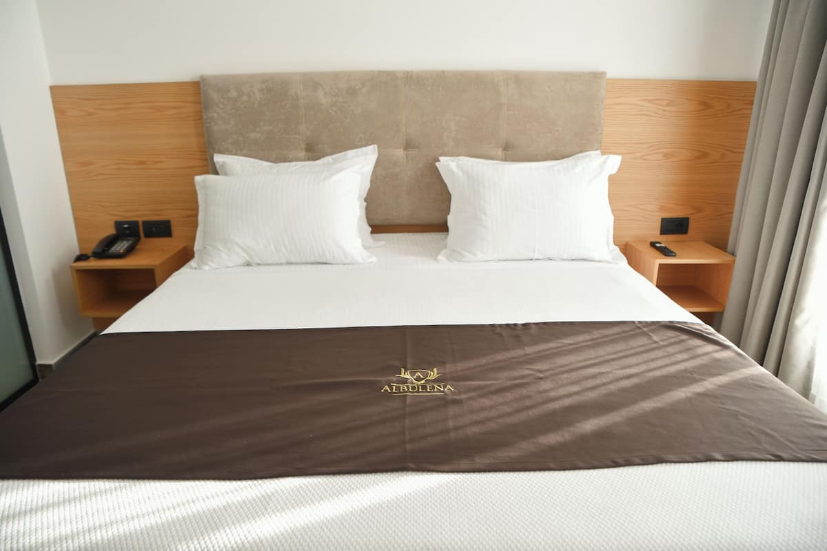 A hotel room with a bed, two pillows, a brown bedspread, wooden headboard, two bedside tables with phones, and a window with curtains.