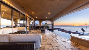 Un espacio de estar al aire libre con techo y vistas al atardecer sobre el océano, con un sofá, sillas y una mesa de comedor.