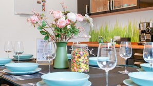 Uma mesa de jantar posta com pratos azul-claros e copos de vinho, um vaso com flores rosa e brancas, e um pote de balas coloridas.