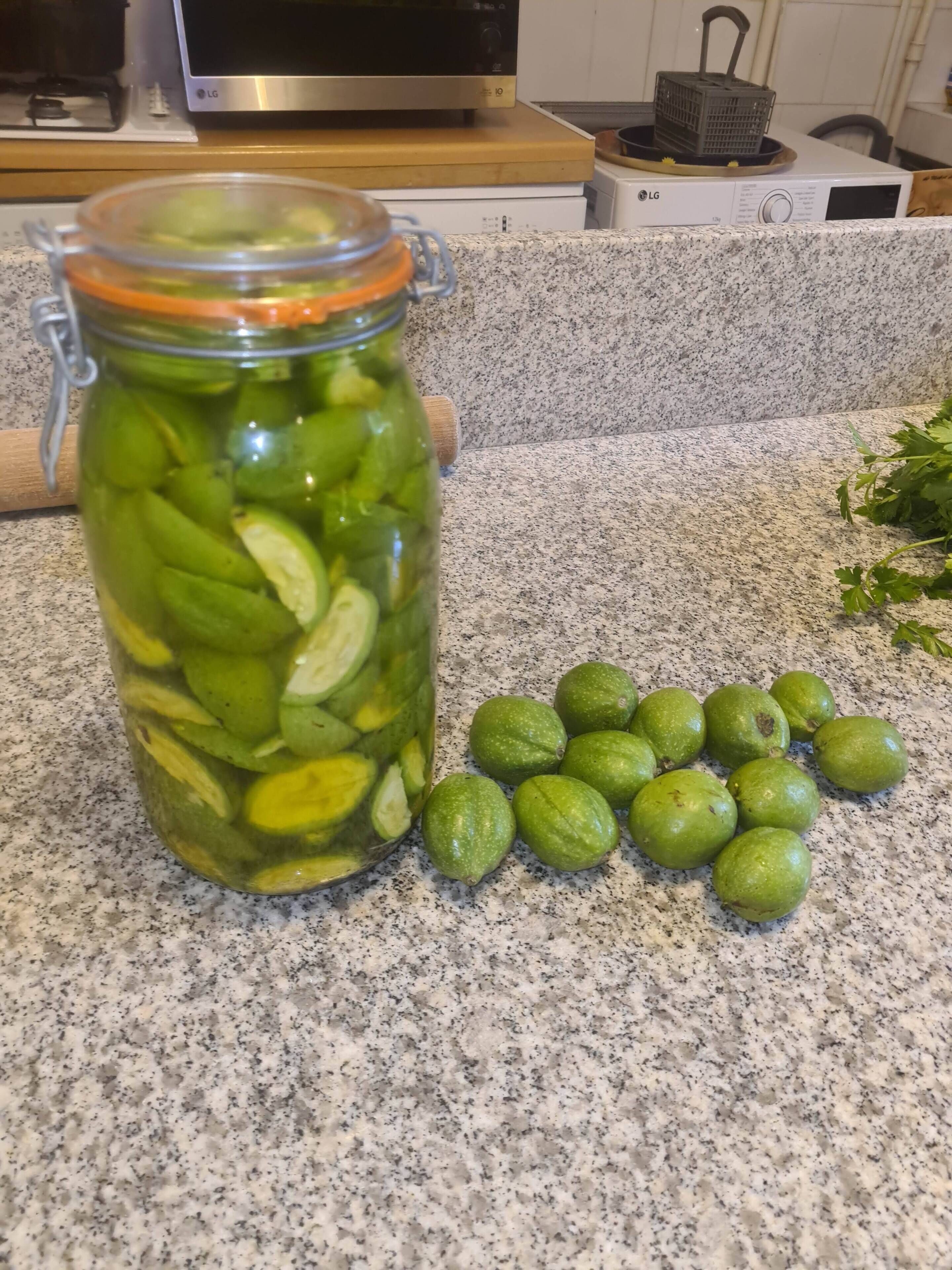 Un comptoir de cuisine sur lequel se trouve un bocal en verre rempli de légumes marinés et de fruits verts entiers.