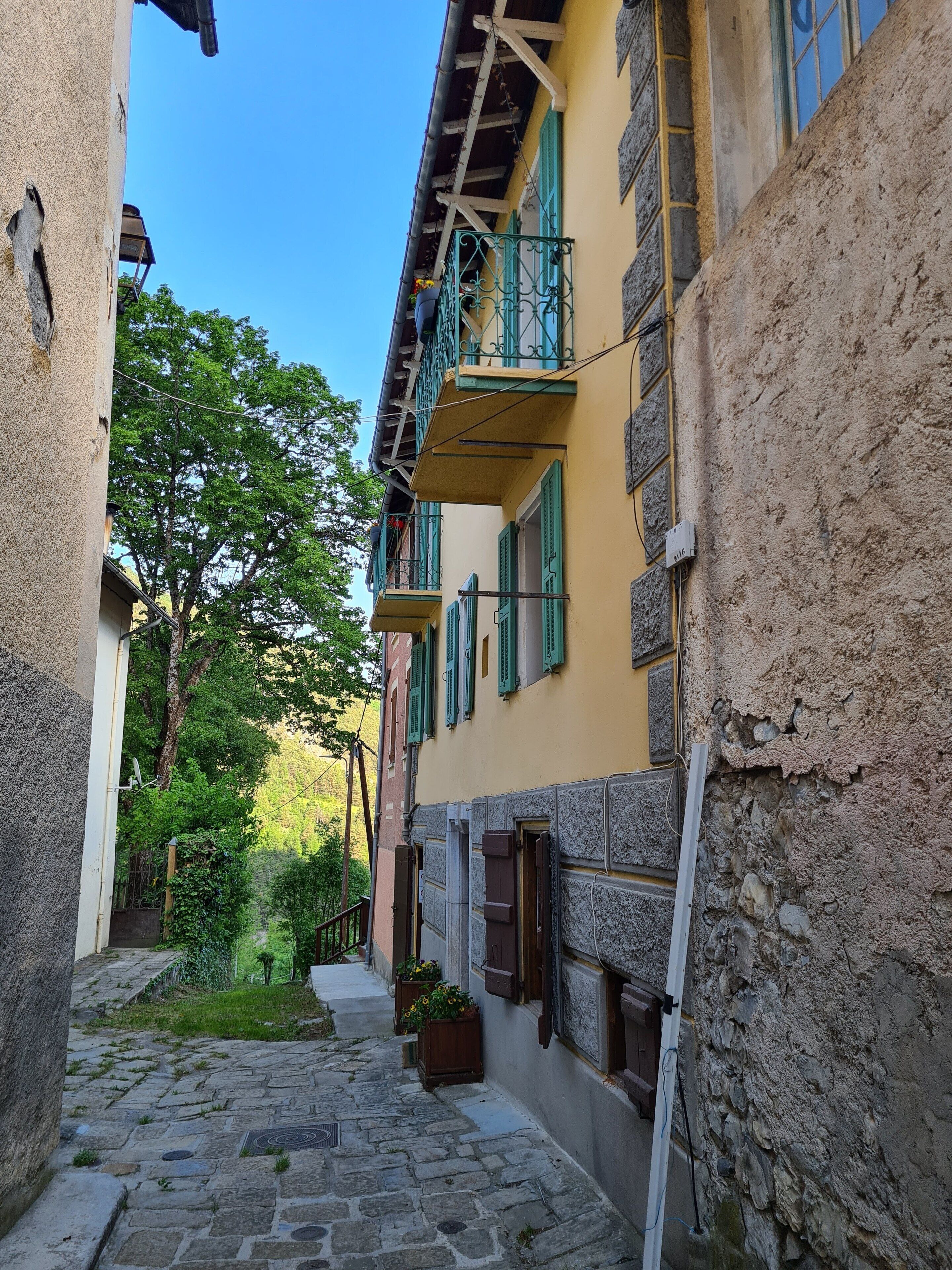 Une ruelle étroite pavée, bordée d’immeubles dotés de balcons et de volets verts.