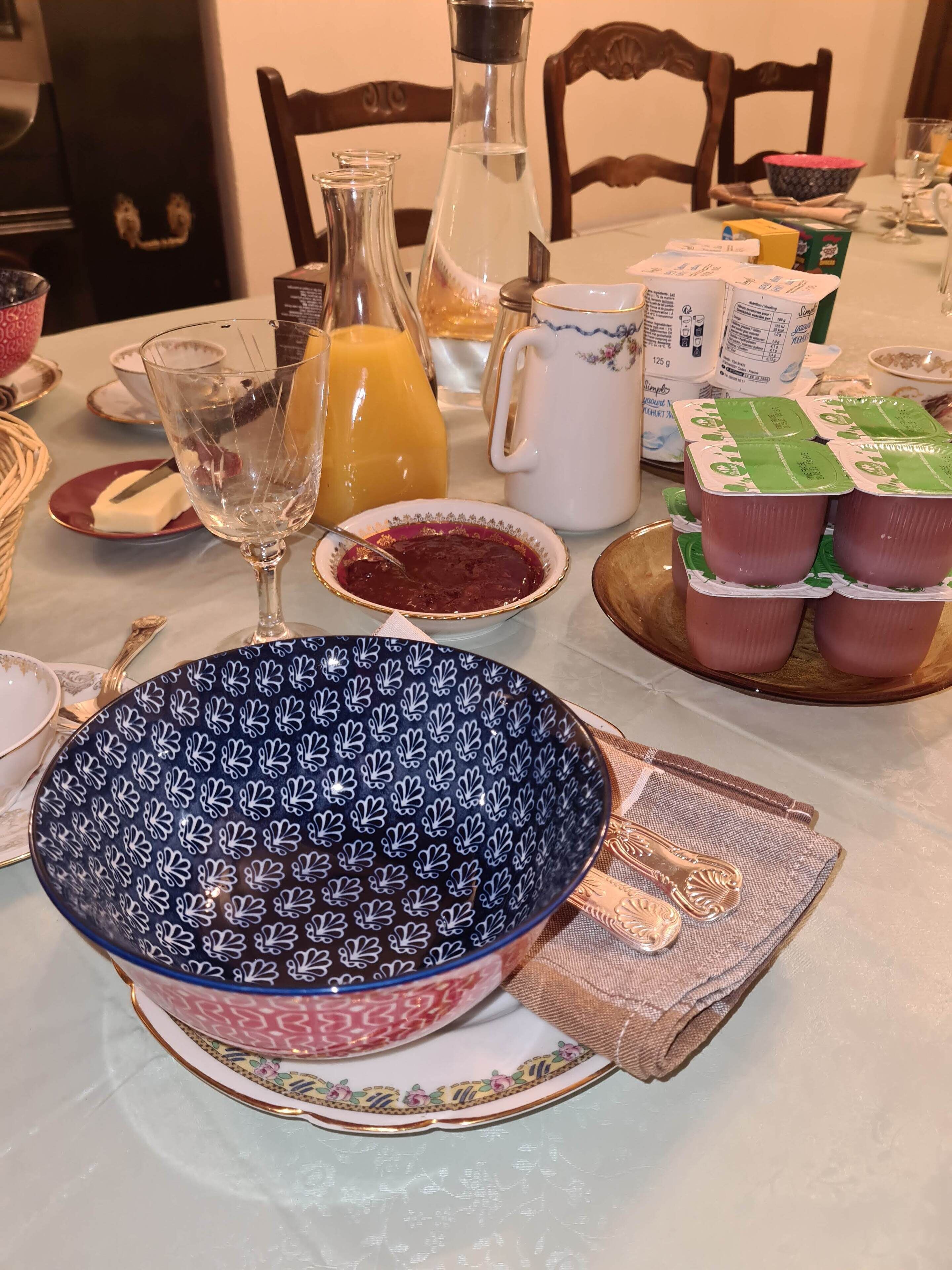 A dining table set with various items including a patterned bowl, a glass pitcher, a jug of orange juice, a white mug, and several packaged food items.