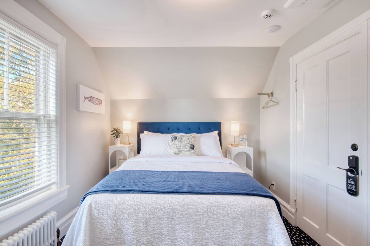 A bedroom with a large bed, white walls, a blue headboard, and a window with blinds.