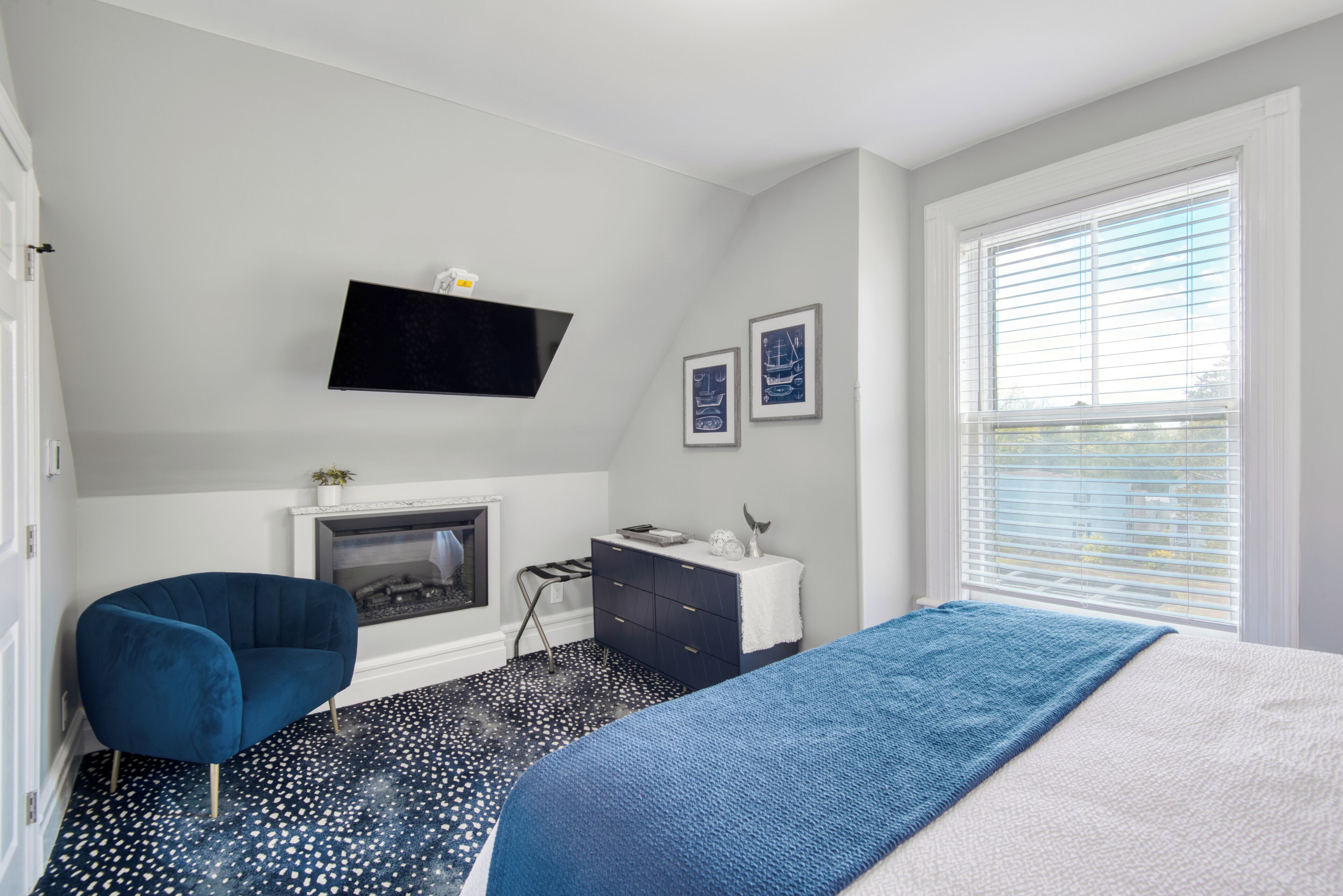 A bedroom with a bed, a blue armchair, a dresser, a flat-screen TV, a fireplace, and a window with blinds.