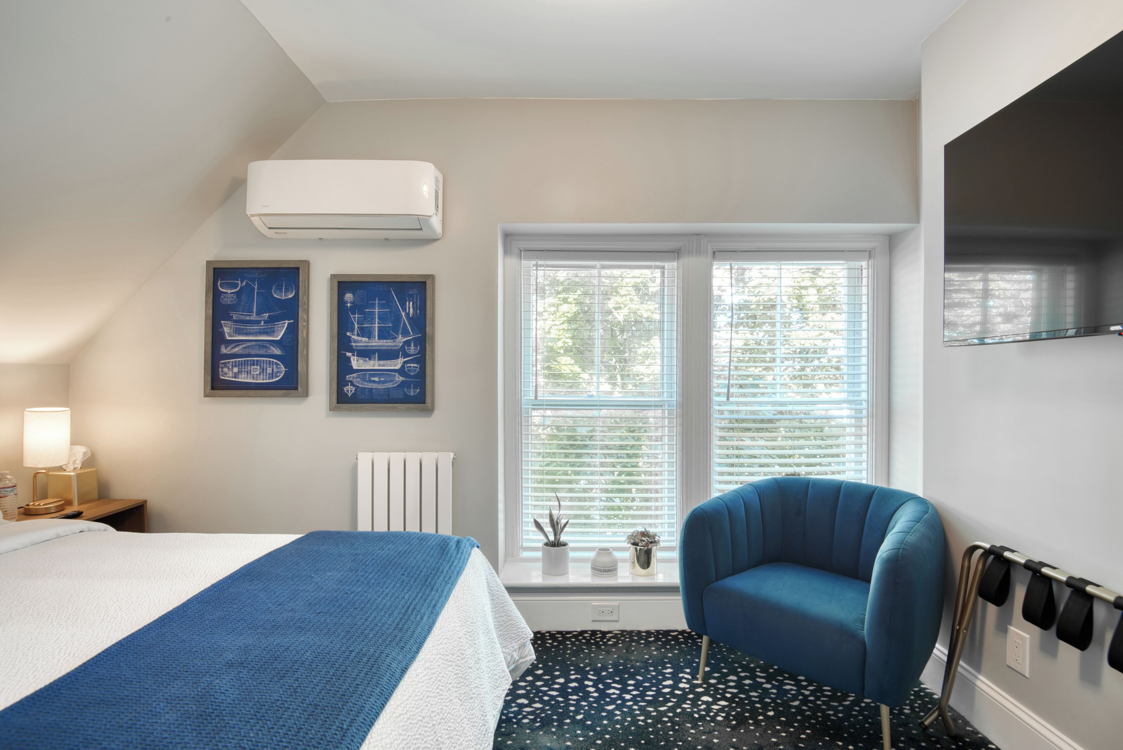 A hotel room with a bed, a blue armchair, a window with blinds, and two framed nautical-themed pictures on the wall.