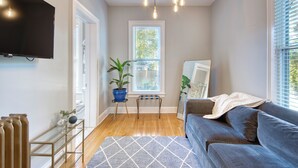 A living room with a blue sofa, a glass coffee table, a TV mounted on the wall, and a window with blinds. - Hudson Navigator (Hudson)