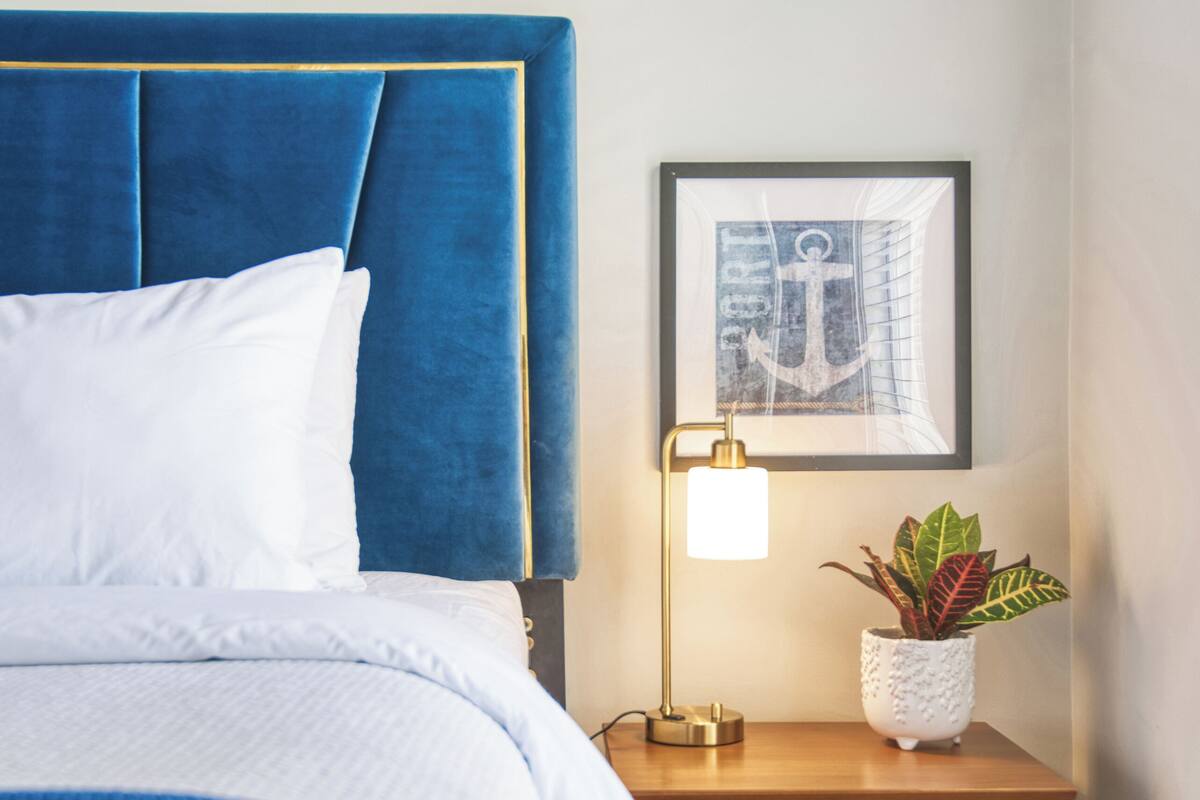 A neatly made bed with a blue headboard, a bedside lamp, a small wooden nightstand, and a framed picture on the wall.