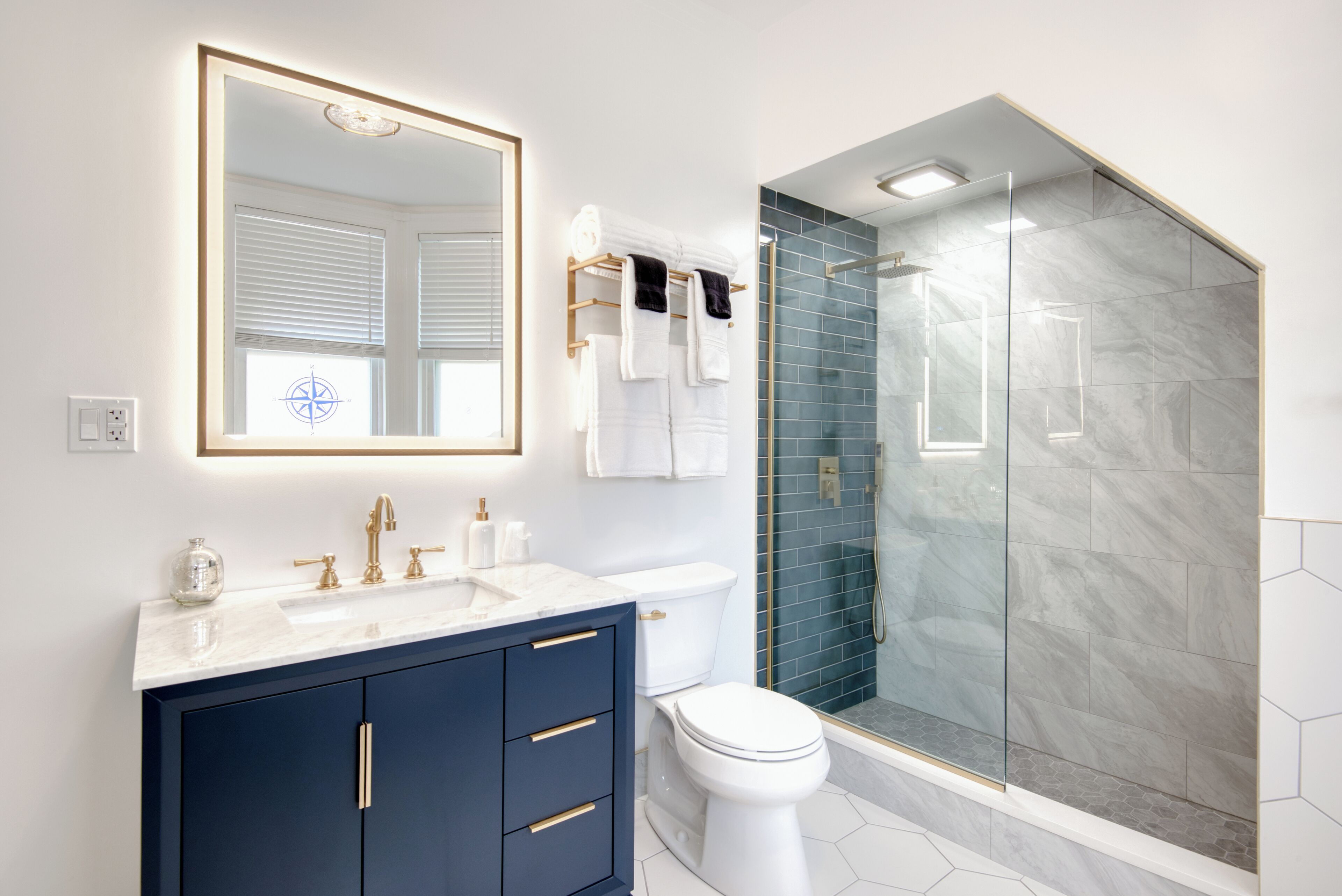 Un baño moderno con ducha envuelta en vidrio, inodoro blanco y tocador azul oscuro con detalles dorados.