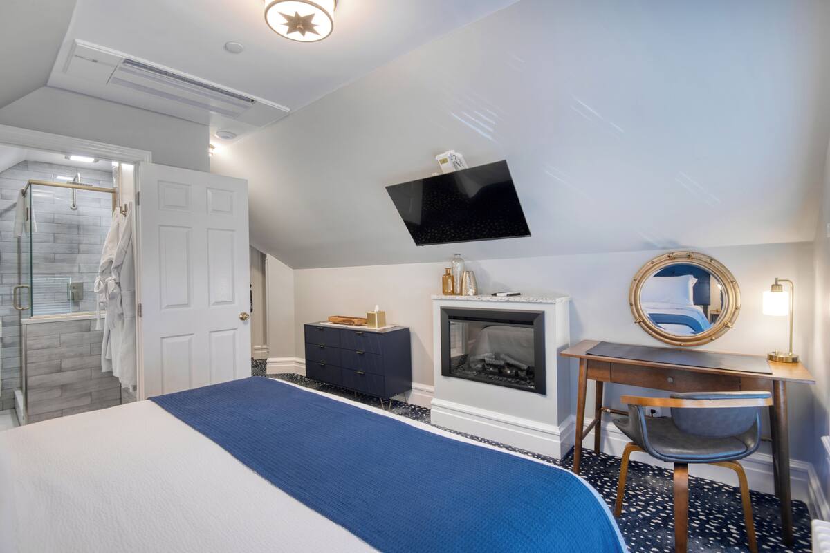 A bedroom with a bed, a desk, a chair, a TV, and a fireplace.