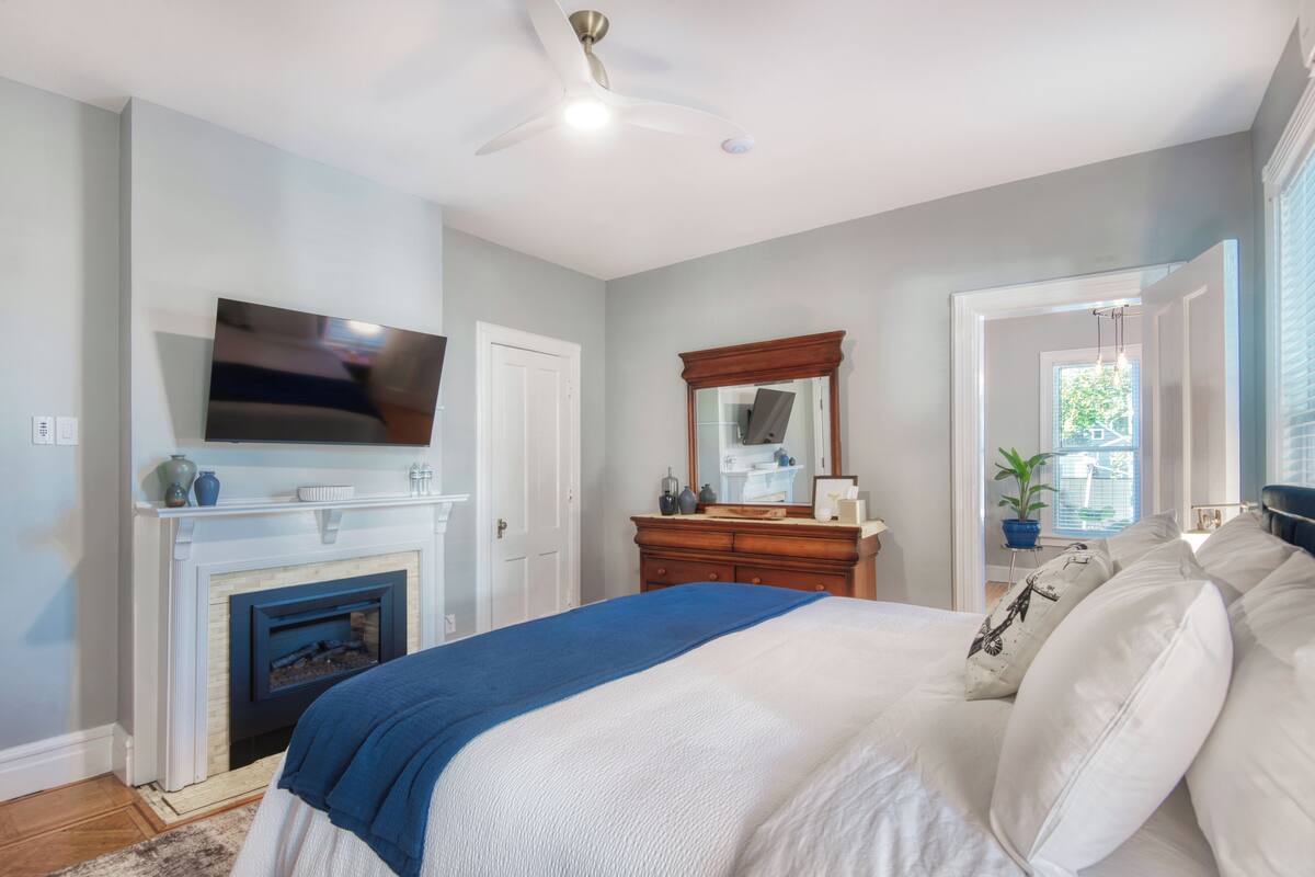 A bedroom with a bed, a television mounted above a fireplace, a dresser, and a mirror.