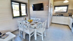 A modern kitchen with white cabinetry, a dining area with white chairs, a TV mounted on the wall, and a window providing natural light. - Holiday Home With Terrace, Sauna (Ustronie Morskie)