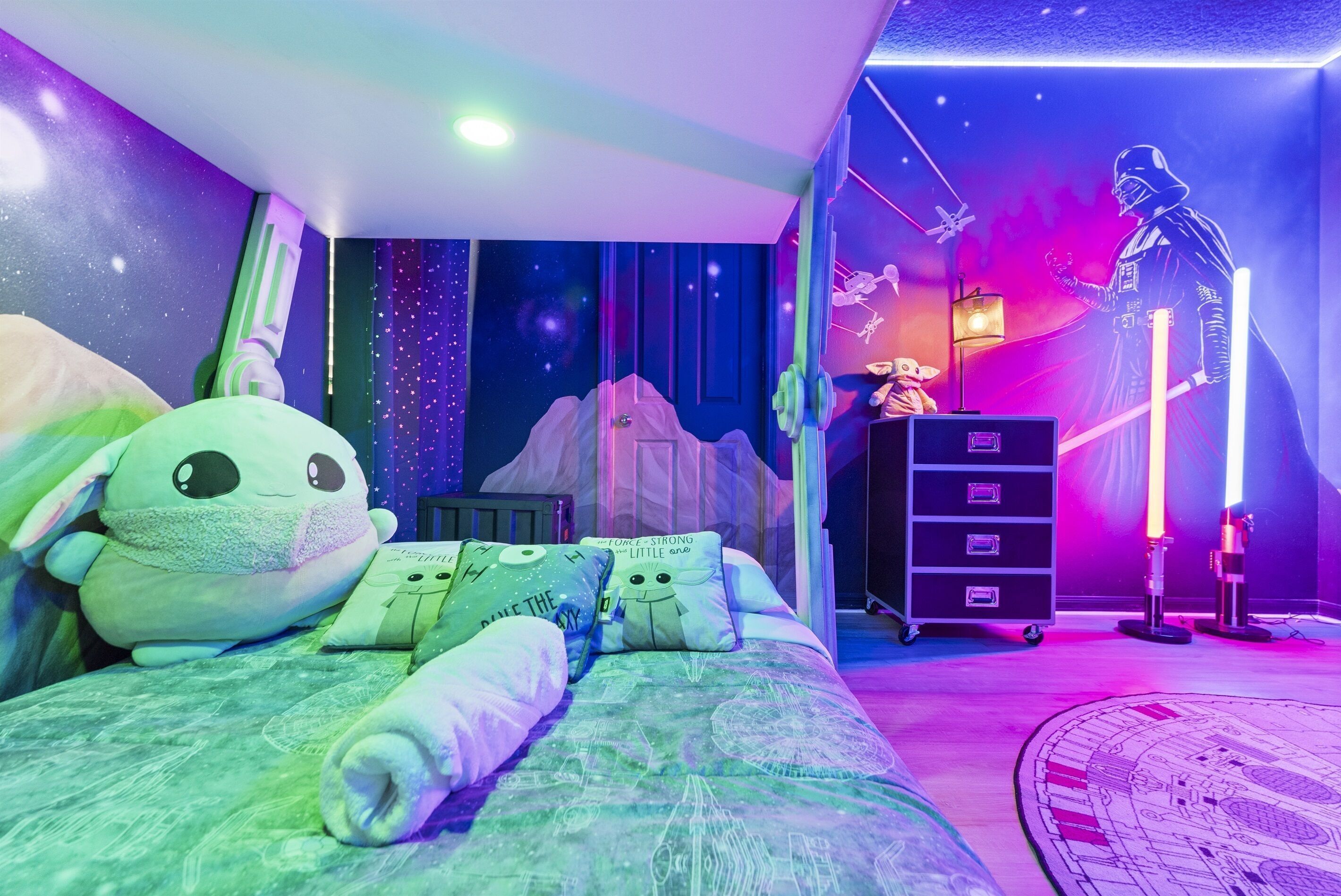 Een kamer met een bed, voorzien van een beddegoed met een sterrenstelselthema, een groot pluche speeltje en kussens met tekst. Er staat een dressoir met lades en een lamp.