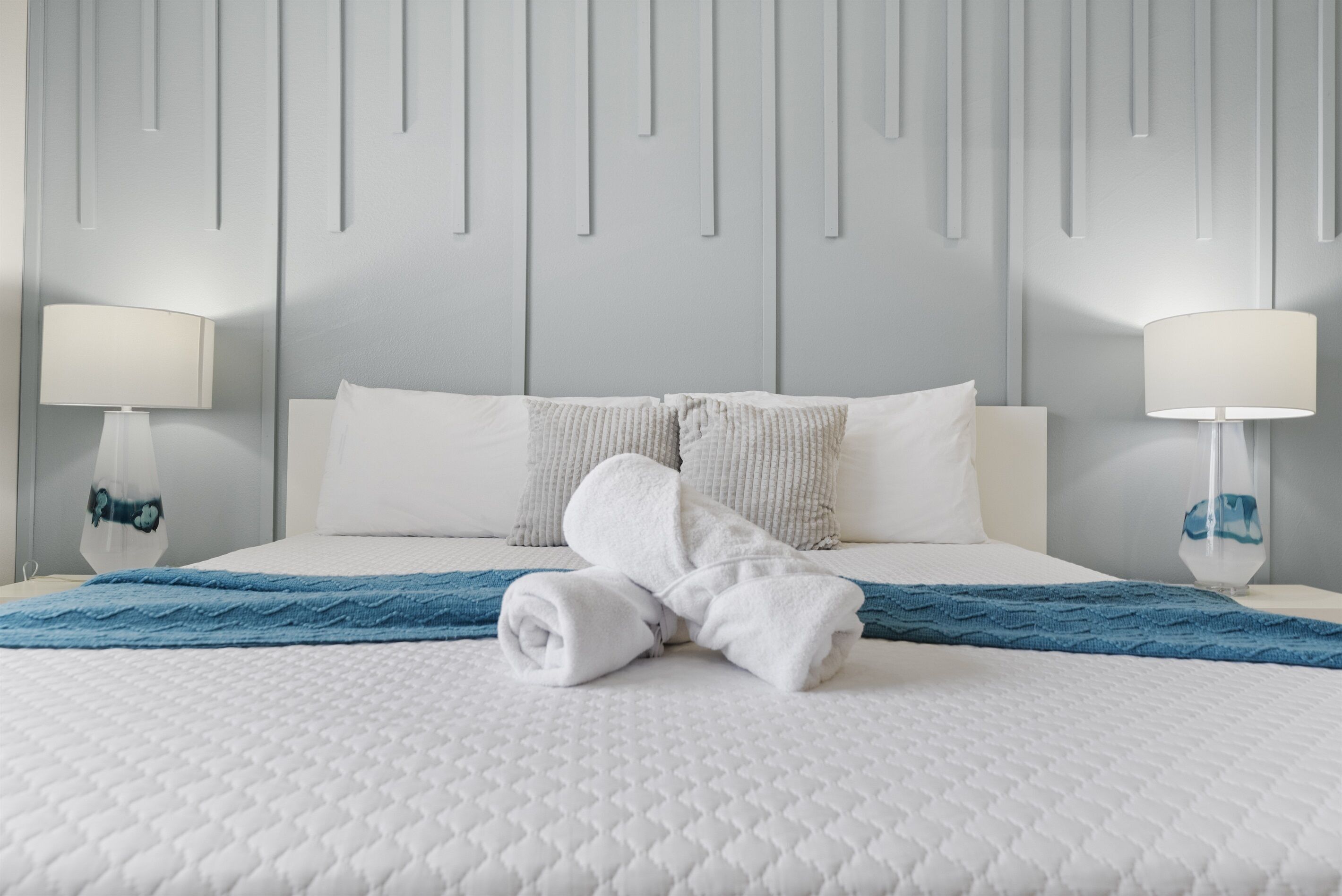 A neatly made bed with white and blue bedding, two bedside lamps, and a wall with vertical lines.