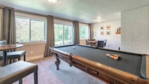 Une table de billard avec des billes, une queue de billard et une chaise, dans une pièce dotée de fenêtres et offrant une vue sur les arbres.