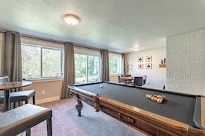 A pool table with balls, a pool cue, and a chair in a room with windows and a view of trees.