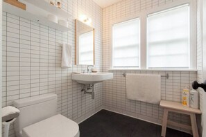 A bathroom with a white tiled wall, a white sink, a toilet, and a window with blinds.
