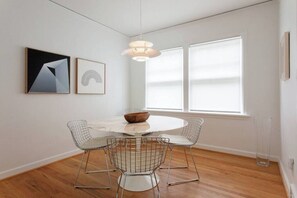 A dining area with a round table, four wireframe chairs, a wooden bowl, and two framed artworks on the wall.