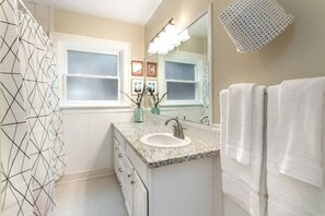 Un baño con lavamanos y tocador blancos, y un espejo grande. Se ve un perchero con toallas blancas y una cortina de ducha con estampado geométrico.