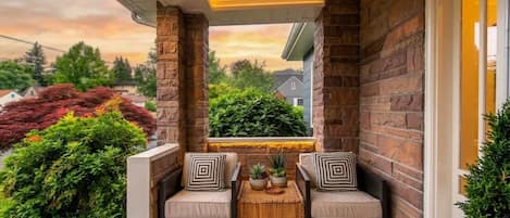 A covered porch with a stone wall, two cushioned chairs, a small table, and a view of the neighborhood.