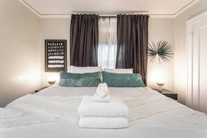 Letto rifatto con lenzuola bianche e un asciugamano piegato al centro. Due comodini con lampade, un quadro appeso al muro e una pianta decorativa a destra.