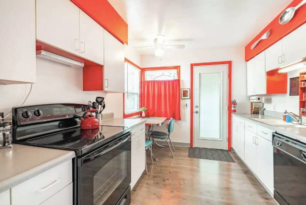 Uma cozinha compacta com armários brancos, uma chaleira vermelha e um espaço de refeições com uma cadeira.