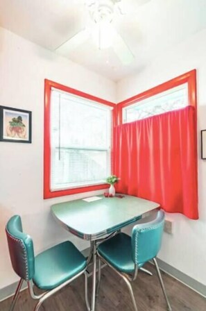 A corner room with a round table and two chairs, a window with red curtains, and a small plant on the windowsill.