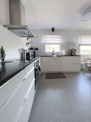 A modern kitchen with white cabinets, a black countertop, and a built-in oven. There are two windows with blinds, a coffee machine, and a sink with a towel hanging on the door.