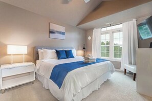 A bedroom with a large bed, white nightstands, a bedside table, a lamp, a window with curtains, and a wall-mounted TV.