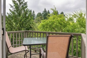 A wooden deck with a glass table and two chairs, surrounded by lush greenery.