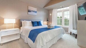 Une chambre à coucher avec un grand lit, deux tables de chevet blanches, une table de nuit, une lampe, une fenêtre avec des rideaux et un téléviseur fixé au mur.