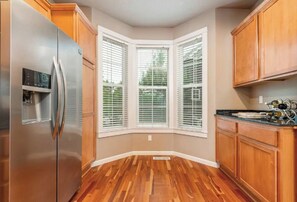 A kitchen with wooden cabinets, a stainless steel refrigerator, and a bay window with white blinds.