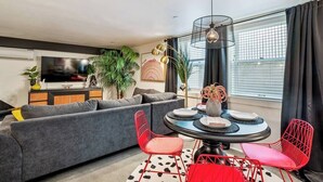 A modern living room with a grey sofa, red chairs, a round table, and a TV on a cabinet. - The Braeburn: Luxury Flat, Steps From NW 23rd! (Portland)